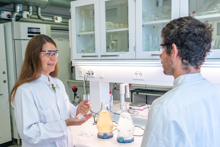 To personer i en laboratorieindstilling, iført kitler og sikkerhedsbriller, står og snakker, mens de arbejder med kemiske reagenser på et bord.