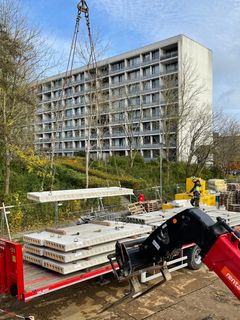 Betonelementer løftes fra en lastbil ved en byggeplads i nærheden af en boligblok.