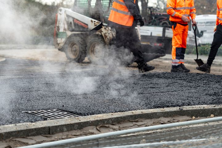 Arbejdere iført orange sikkerhedstøj står ved nyudlagt asfalt, hvor damp stiger op. En lille arbejdsbil ses i baggrunden.