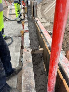 Beton bliver hældt i en form til en støttemur ved motorvejsudvidelse. Arbejdere ses i baggrunden.