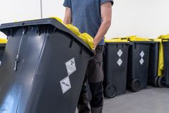 En person trækker en mørkegrå affaldscontainer med gult låg og farlige gods symboler.