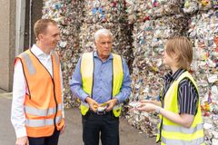 Tre personer i sikkerhedsveste står foran stablede plastballer og diskuterer genanvendelse.
