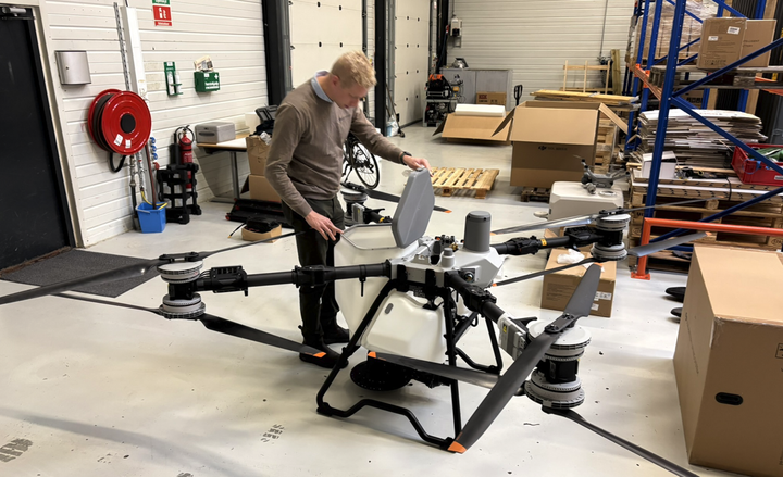 En mand justerer en stor landbrugsdrone med sprøjtetank i et værksted med reoler og papkasser i baggrunden.