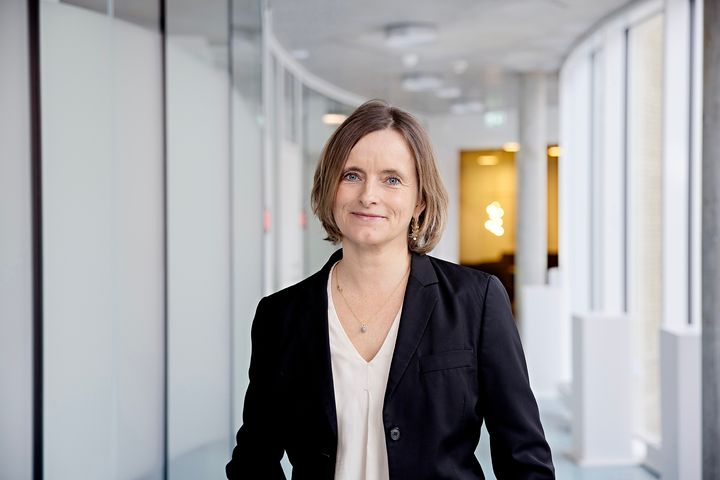 Smilende kvinde med lyst pagehår i sort blazer står foran utydelig baggrund af buet gang