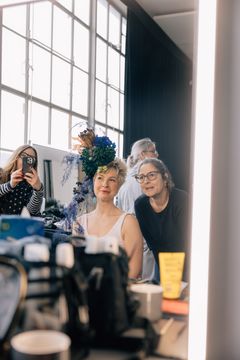 En spinkel lyshåret kvinde sidder ved et spejl / sminkebord og bliver stylet af to andre kvinder. En arbejder med makeup, en anden med blomster til den lyshårede kvindes hår. Der er et meget stort vindue i baggrunden.