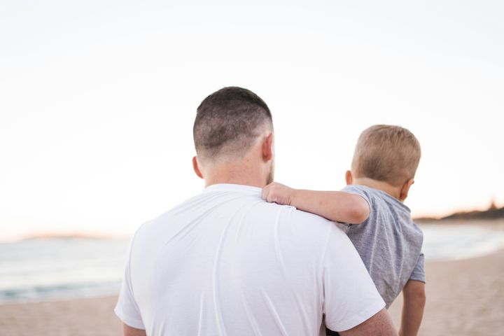 Far bærer sin tre-årige søn, de har ryggen til og går på stranden