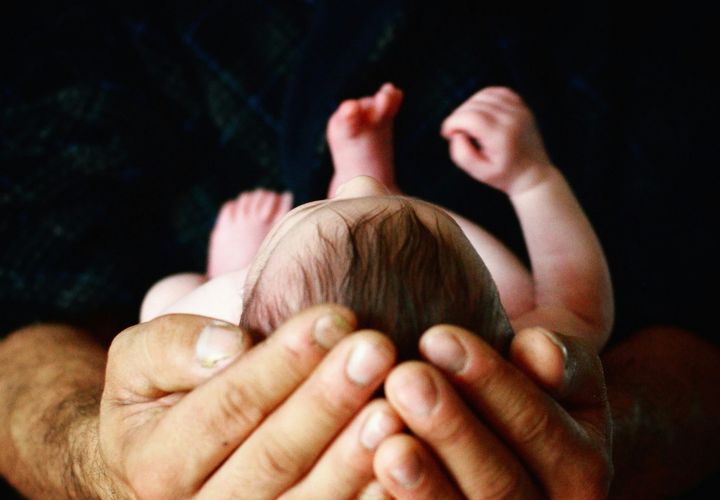 Farvefoto af en baby, der ligger på ryggen i en mands hænder. Babyen har mørkt hår. Hænderne har slidte negle