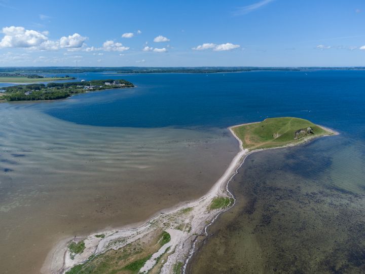 Luftfoto af en kystlinje med klart blåt vand og grønne områder under en skyfri himmel.