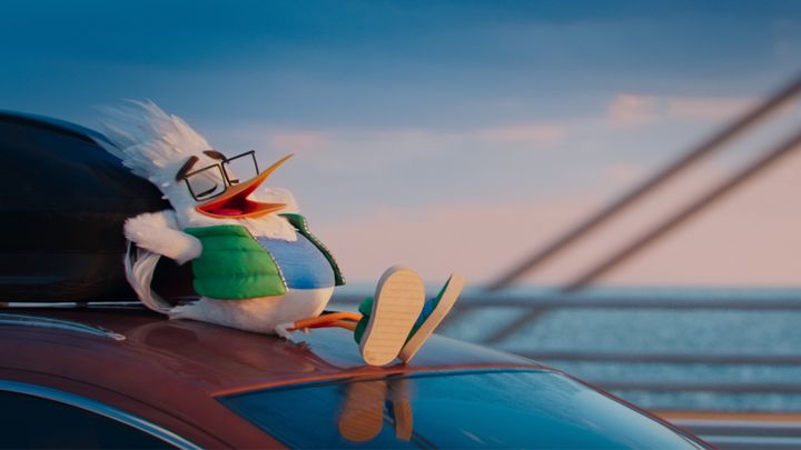 En animeret fugl med briller og en grøn vest sidder afslappet oven på en bil mod en baggrund af hav og bro.