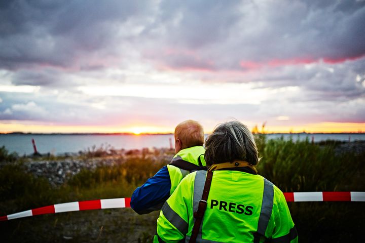 To personer iført presseveste står ved en afspærring og ser ud over vandet ved solnedgang.