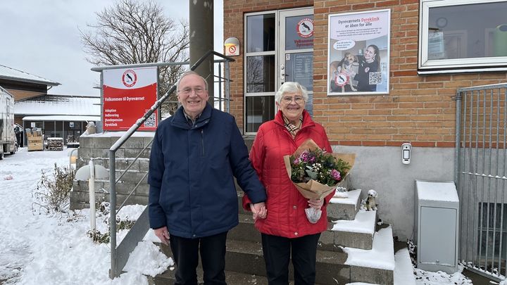 Billede af Joan og Bendt Refsgaard stående sammen foran hovedindgangen til Dyreværnet i Rødovre. Joan (til højre) har en stor buket blomter i hånden. Der ligger sne i gården i baggrunden.
