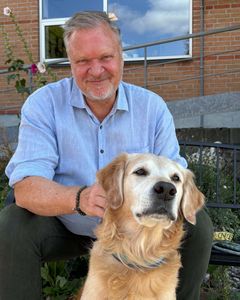 En mand sidder på en bænk udenfor en bygning sammen med en golden retriever.