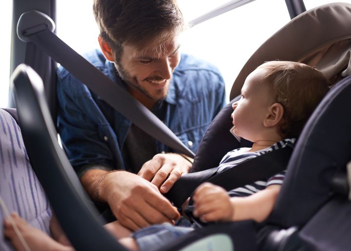 Et barn sidder i en autostol, mens barnets far smiler til barnet.