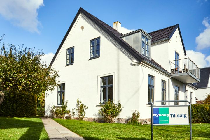 Hvid villa med græsplæne og "Til salg"-skilt fra Nybolig i forgrunden.