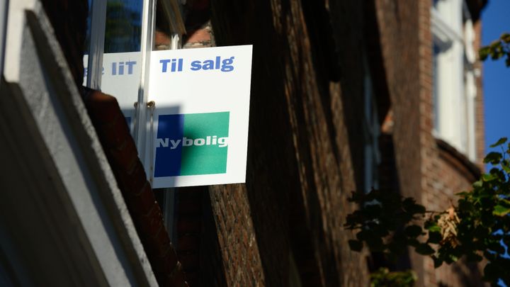 Skilt med teksten "Til salg" fra Nybolig hænger ud af et vindue på en bygning.