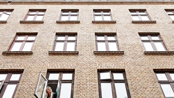 En person kigger ud af et åbent vindue i en murstensbygning med mange vinduer.