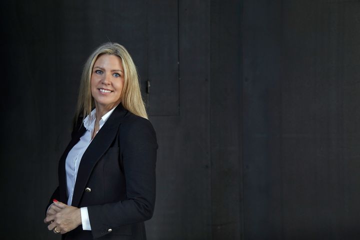 Jeg ved ikke, hvem personen er, men billedet viser en kvinde i forretningsdragt, der smiler og står foran en mørk baggrund.