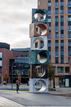 En høj skulptur af stablede stenklodser med runde udskæringer står udendørs foran VIA i Horsens.