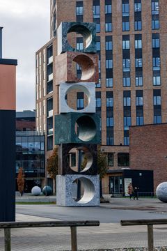 En høj skulptur af stablede stenklodser med runde udskæringer står udendørs foran VIA i Horsens.